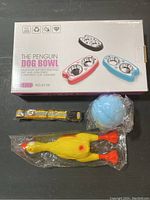 Dog essentials bundle showing boxed silicone dog bowl, medium yellow reflective collar, and blue rubber squeaky chicken dog toy in packaging.