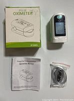 Pulse oximeter device with its original packaging and additional accessories (manual, USB cable) laid out.