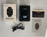 Complete lot contents showing the smart watch in black silicone band seated in black foam insert, original white box with smart watch image and specs, charging cable, warranty card, and user manual.