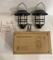 Two black solar wall lanterns with mounting hardware and instruction sheet, and the product box labeled 'Solar Wall Lantern 2 Pack'.