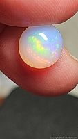Close-up of a round cabochon Ethiopian opal gemstone held between fingers, showcasing its vibrant pastel color flash and smooth polished surface.