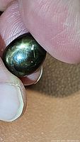 Close-up photo of a person's fingers holding a large, dark black-green cabochon sapphire displaying a star effect on its surface.