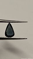 Pear-shaped blue green sapphire 0.79 carat held in tweezers against neutral background, showing color and clarity.