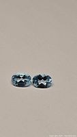 Pair of oval cut natural sky blue topaz gemstones laying side by side on a neutral background, showing their size and color clearly.