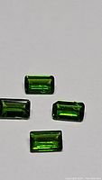 Four rectangular step-cut electric green Russian chrome diopside gemstones approx. 5x3mm each with a total weight of 1.16ct shown on plain background.