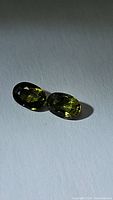 Two oval cut multicolored sapphires with green and yellow tones shown on a white background with shadow effects.