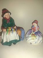 Two Royal Doulton porcelain figurines: one elderly woman sewing ribbons, another young woman with a basket of flowers.