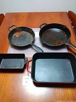 All four pieces of cast iron cookware on wooden table: two round skillets and two rectangular pans, one with red handles, showing surface rust and wear.