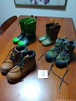 Four pairs of children's boots on wooden table: Minecraft rain boots, dump truck rain boots, green hiking boots, and brown hiking boots. Includes lot sticker number 67.