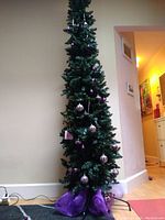 Full view of the tall, narrow Christmas tree showing pencil style shape and overall decoration with purple and pink striped ornaments and purple mesh around the base.