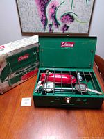 Image showing green Coleman two-burner camping stove open with metal grill, red fuel tank, two circular burners, and original box on the side.