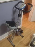 Foldable exercise bike standing on a wooden floor with a black cushioned seat, handlebar with control/display panel, and pedals.