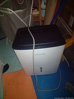 Frigidaire branded white and navy blue dehumidifier with power cord, placed on a floor with other household items surrounding it.