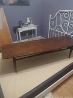 Rectangular mid-century modern wooden coffee table with tapered legs, visible signs of wear and age on surface, placed in a room with a white metal bed frame and small side table.