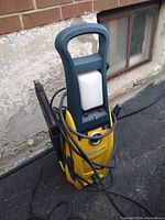 Front side view of yellow and blue electric power washer with white detergent bottle and handled spray gun wrapped with hose cords.