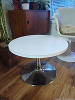 Round white tabletop with chrome base shown from side angle with nearby furniture and wood floor.