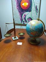 Balance scale and blue globe placed on wooden table with artwork in the background.