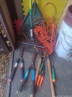 Photo showing two rakes, three garden pruners/shears, and an orange coiled extension power cord against a yellow wall background