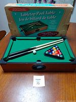 Full view of tabletop pool table set including the table with green felt surface, two cues, white ball, and colored balls in rack alongside the original box.