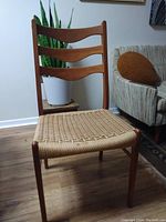 Front view of teak dining chair with three curved horizontal slats on backrest and woven rattan seat on wood floor next to couch and plant.