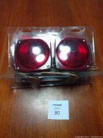 Front view of two rectangular red trailer tail lights in plastic packaging on wooden surface, showing clear mounting holes and red lenses.