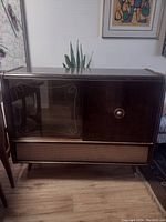 Full front view of vintage bar radio cabinet showing wooden finish, glass panel on left, door with handle on right, and speaker grille below.