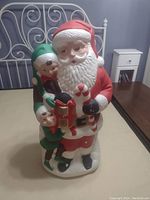 Photo showing vintage Santa figurine approximately 30x18 cm with two attached elves, displays color and detail.