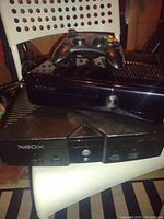 Photo showing both the Xbox 360 console with an Xbox 360 controller on top and the original Xbox console placed beneath. Consoles and controller are black with visible signs of use.