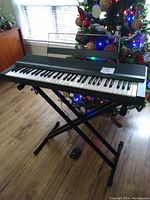 Yamaha keyboard on black adjustable X-style metal stand with foot pedal on the wooden floor near a Christmas tree.