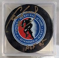 Top view of black hockey puck showing the Hockey Hall of Fame logo and Cam Neely autograph in gold marker with 'HOF 05' inscription.