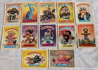 Photo showing 12 Garbage Pail Kids cards laid out on a flat surface, displaying characters and card titles clearly.