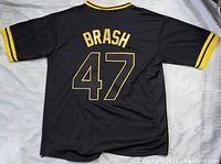 Back of black jersey showing 'BRASH' and number '47' in large yellow outlined font with yellow triple stripe on sleeve cuffs.