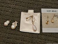Two pairs of pearl earrings and one 14k gold pearl pendant shown on a white velvet card. One pair of earrings has baroque pearls, the other pair has a pearl and small stones cross design.