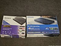 Photo showing both Sony BDP-BX650 and Samsung BD-J6300 Blu-ray player boxes side by side on carpeted floor.
