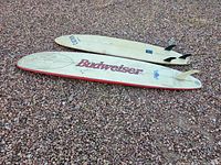 Two full-size surfboards on gravel ground, one with Budweiser logo and red trim, the other plain white showing wax spots and fins.
