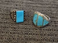 Two sterling silver turquoise rings placed on textured dark surface, showing rectangular stone and inlaid design.