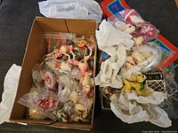 Photo showing an open box filled with multiple vintage Christmas ornaments, some loose and some wrapped in plastic, alongside other decorations wrapped in tissue paper.