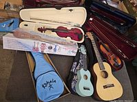 Wide view showing four ukuleles including one boxed new Kohaia ukulele with accessories, next to three violins in open cases