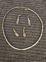 Tri-color sterling silver necklace laid flat with lobster clasp visible. Two pairs of dangle earrings included: one tri-color matching feather design, one silver-toned feather design. All items displayed on textured brown surface.