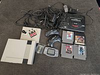 Overview of vintage video game lot including Nintendo NES console, Sega Genesis console, Game Boy Advance, controllers, cables, and game cartridges spread on a dark fabric surface.