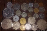 Photo of a collection of approximately 24 older coins from various countries, laid out on a wood surface, showing assorted designs, sizes, and materials