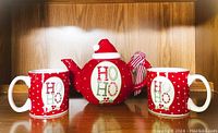 Photo displaying teapot with two mugs on wooden shelf, showing front view with 'HO HO' text and Christmas theme design.