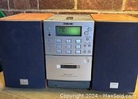 Front view of Sony PMC-D307L main unit flanked by left and right speakers on either side, showing digital display tuned to FM station and control buttons.