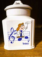 Front side of white ceramic canister showing dancing Tetley Tea Folk character and musical notes in blue and cartoon colors