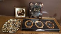 Photo showing all items in the lot displayed together on a wooden surface, including the ceramic picture, metal tray, mirror, and the boxed set of Bombay photo frames