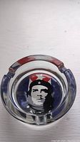 Top view of clear round glass ashtray with a black and white image of Che Guevara in the center and a red, white, and blue patterned background.