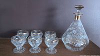 Set of MCM clear glass decanter and six matching liqueur glasses with white floral and gold painted designs on a dark background