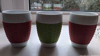 Front view of the set of three Bodum Pavina mugs with 2 red and 1 green silicone sleeve, showing the textured surfaces and cylindrical shape.