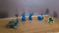 Five small animal figurines arranged on a surface: blue glass teddy bear, elephant (tail chipped), swan, frog, and a green ceramic frog.