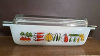 Side view of white oblong casserole with colorful vegetable pattern, clear glass lid on top.
